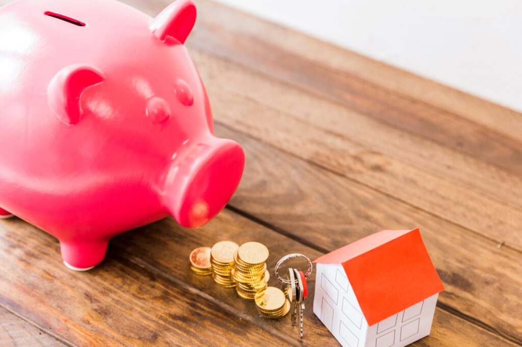 Piggybank, gold coins, keys, and miniature house on a wooden desk in the concept of 'how to save money when moving to Balham'.