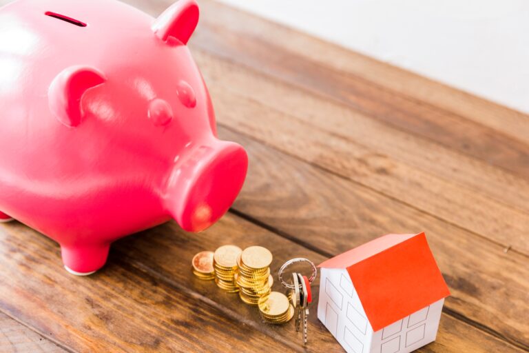 Piggybank, gold coins, keys, and miniature house on a wooden desk in the concept of 'how to save money when moving to Balham'.